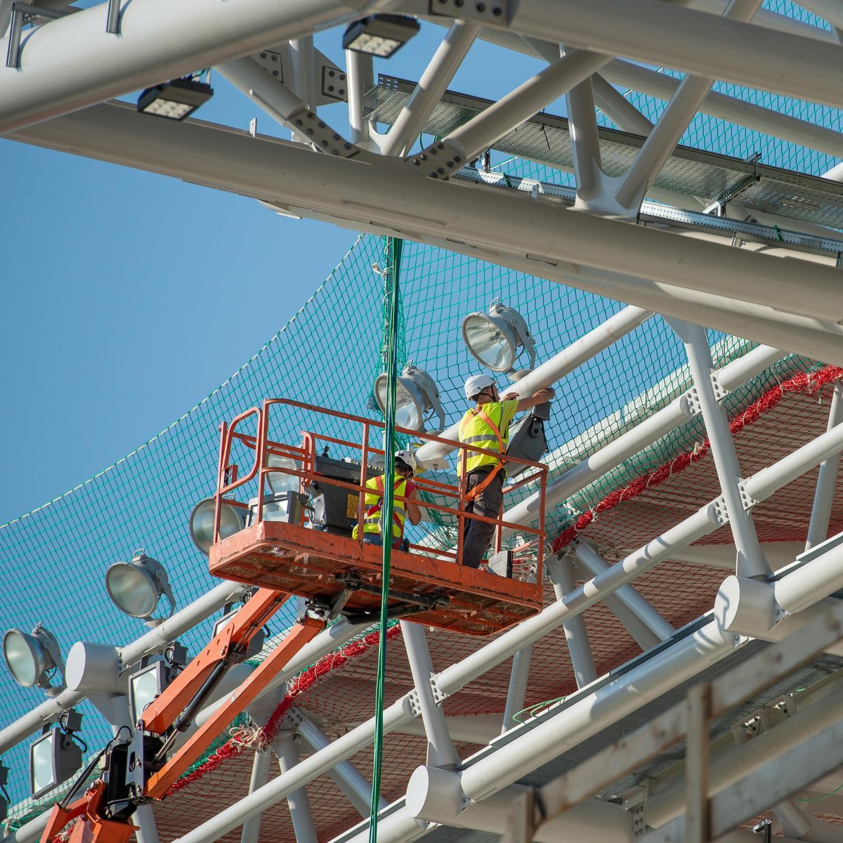 Worker in yellow safety vest on elevated hydraulic lift platform against clear blue sky and industrial building structure